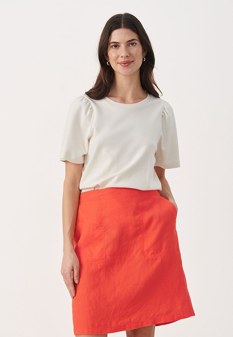 Kurzärmlige cremefarbene Bluse mit gepufften Schultern, kombiniert mit einem leuchtend orangen A-Linien-Rock mit Taschen und strukturiertem Stoff.