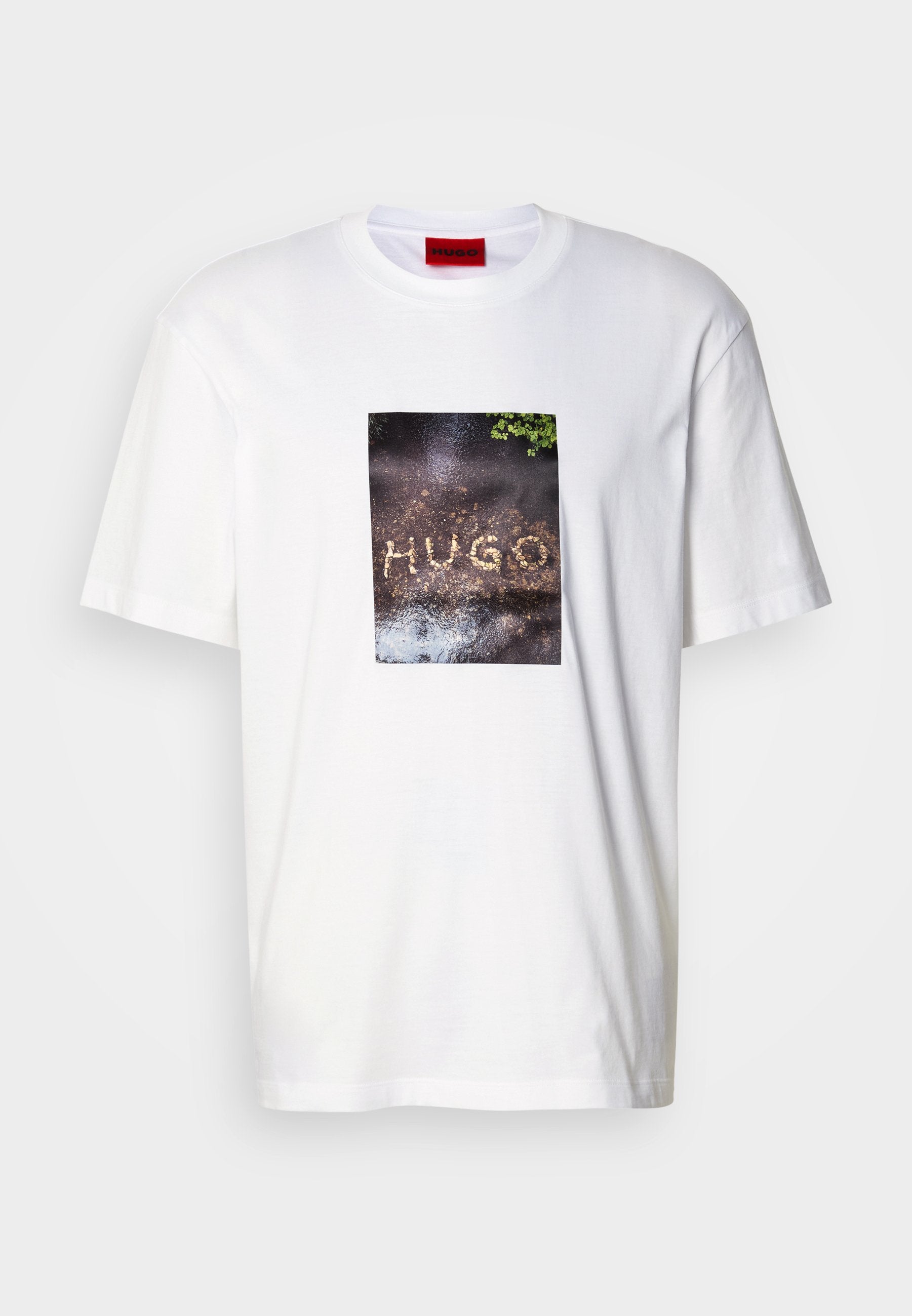 Weißes Baumwoll-T-Shirt mit einem rechteckigen Grafikdruck und dem Schriftzug "HUGO" auf einer strukturierten, dunklen Oberfläche, umgeben von Grünpflanzen.