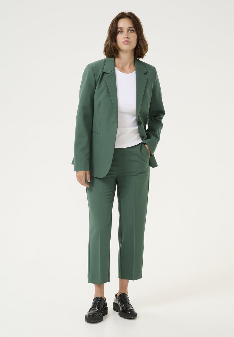 Grüner maßgeschneiderter Anzug mit einem einreihigen Blazer und verkürzten Hosen, getragen über einem weißen Oberteil. Schwarze Schuhe vervollständigen das Ensemble.