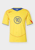 Gelbes Sporttrikot mit blauem Kragen, roten Paspelierungen, rotem Nike-Logo, roten Ärmeldetails und einer blauen "90" in einem Kreis auf der Brust.