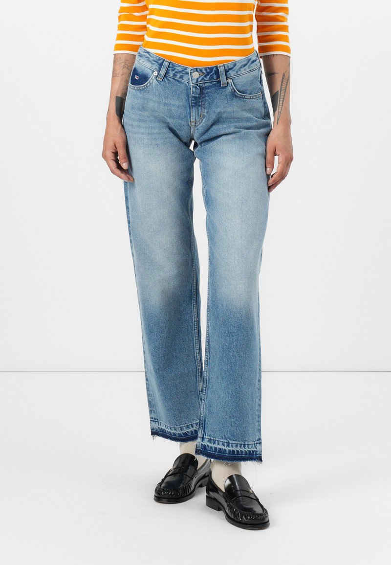 Person, der hellblaue weite Jeans mit rohen Säumen, schwarze Slipper, weiße Socken und ein orange-weißer gestreifter Oberteil trägt.