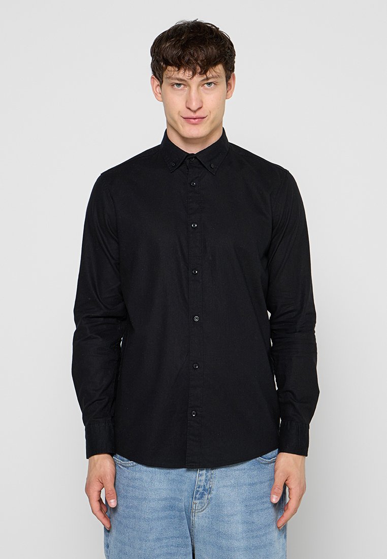 Junger Mann, der ein schwarzes Hemd mit Knöpfen und hellblaue Jeans trägt, steht vor einem einfarbigen hellgrauen Hintergrund.