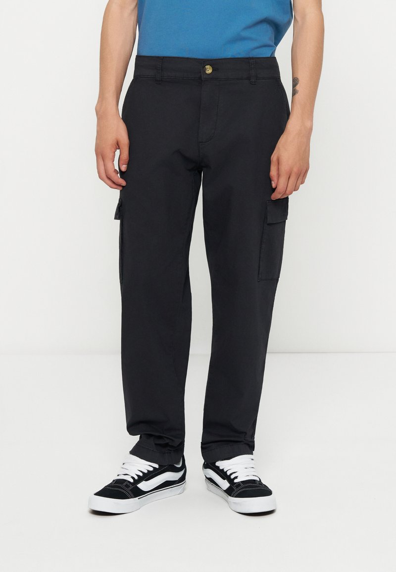 Mann trägt schwarze Cargohosen mit Seitentaschen, schwarz-weiße Sneakers und ein blaues Hemd, steht vor weißem Hintergrund.