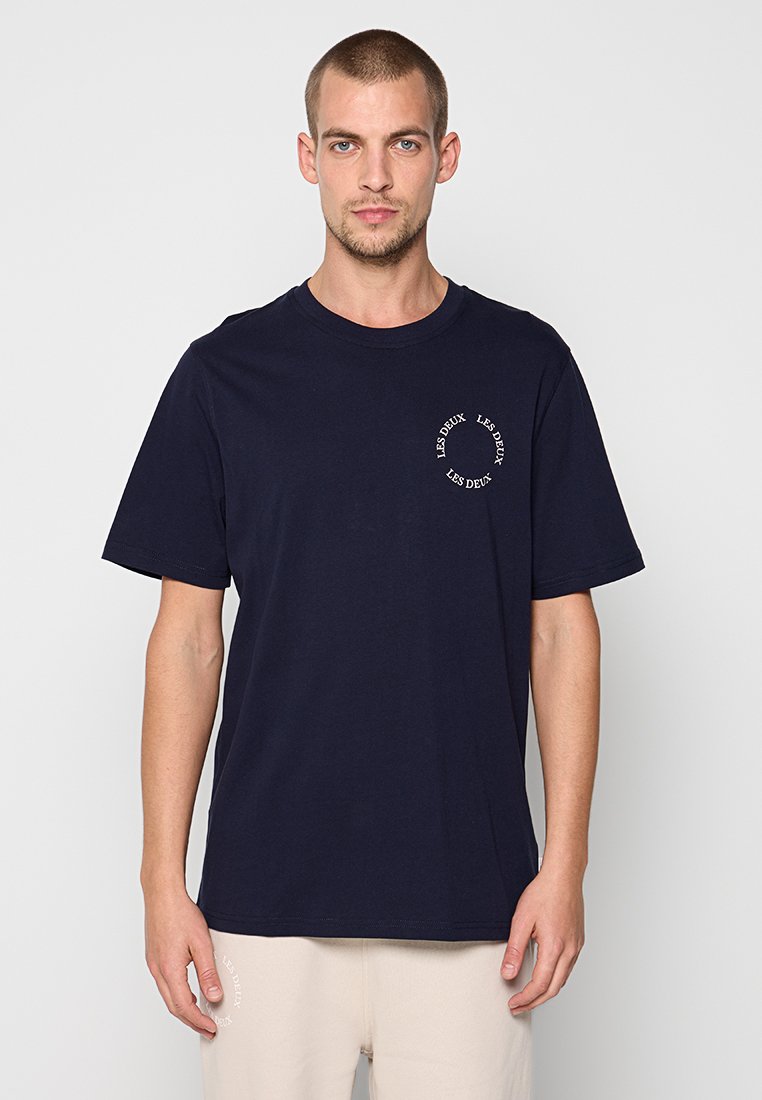Marineblaue Baumwoll-T-Shirt mit Rundhalsausschnitt und kurzen Ärmeln; mit kreisförmigem weißen Text im linken Brustbereich. Glatte Stoffstruktur.