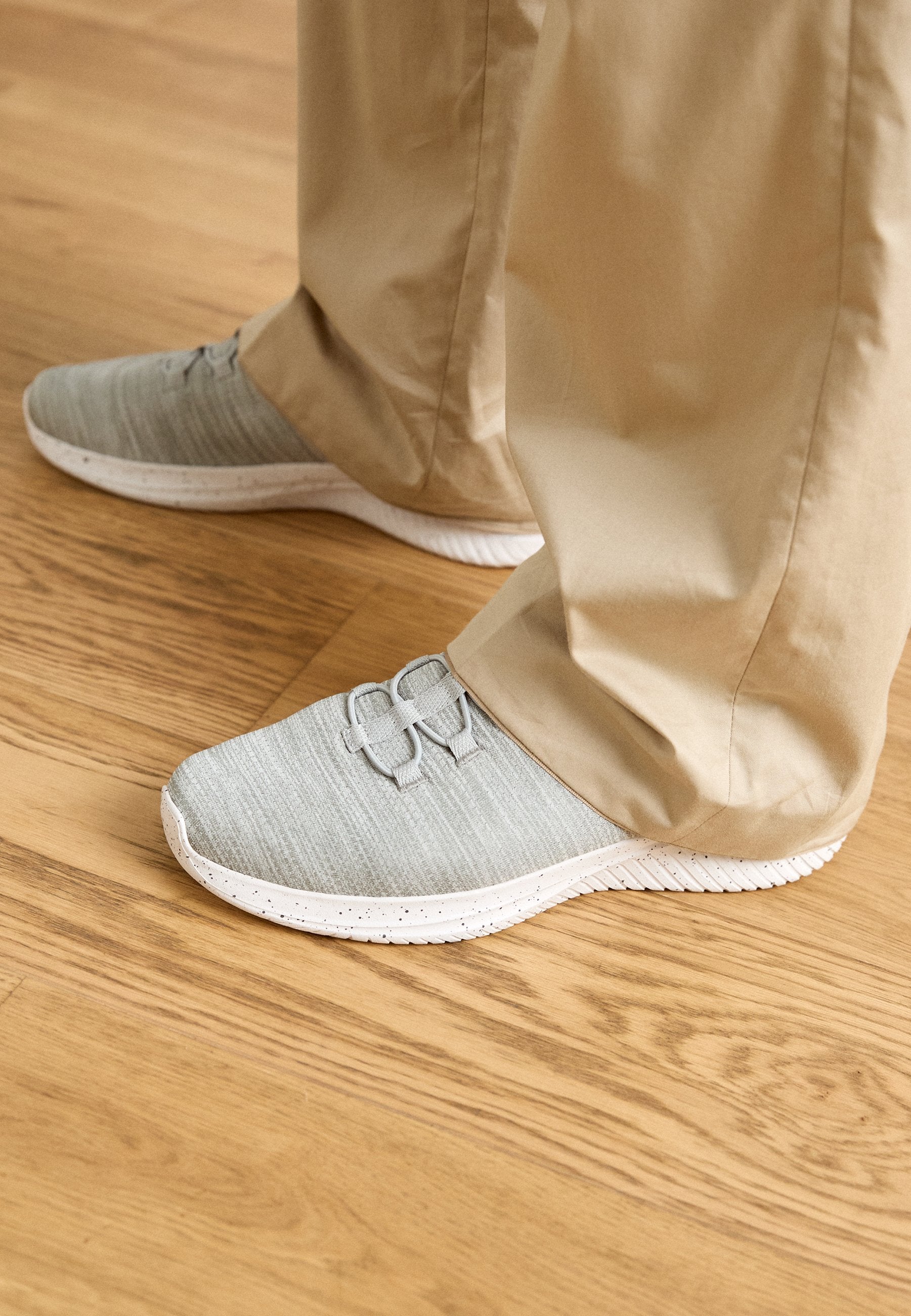Helle graue Sportschuhe mit einem strukturierten Obermaterial und einer weiß gesprenkelten Sohle, versehen mit dekorativen Schnürsenkelakzenten. Tragen Sie sie mit beigen Hosen.