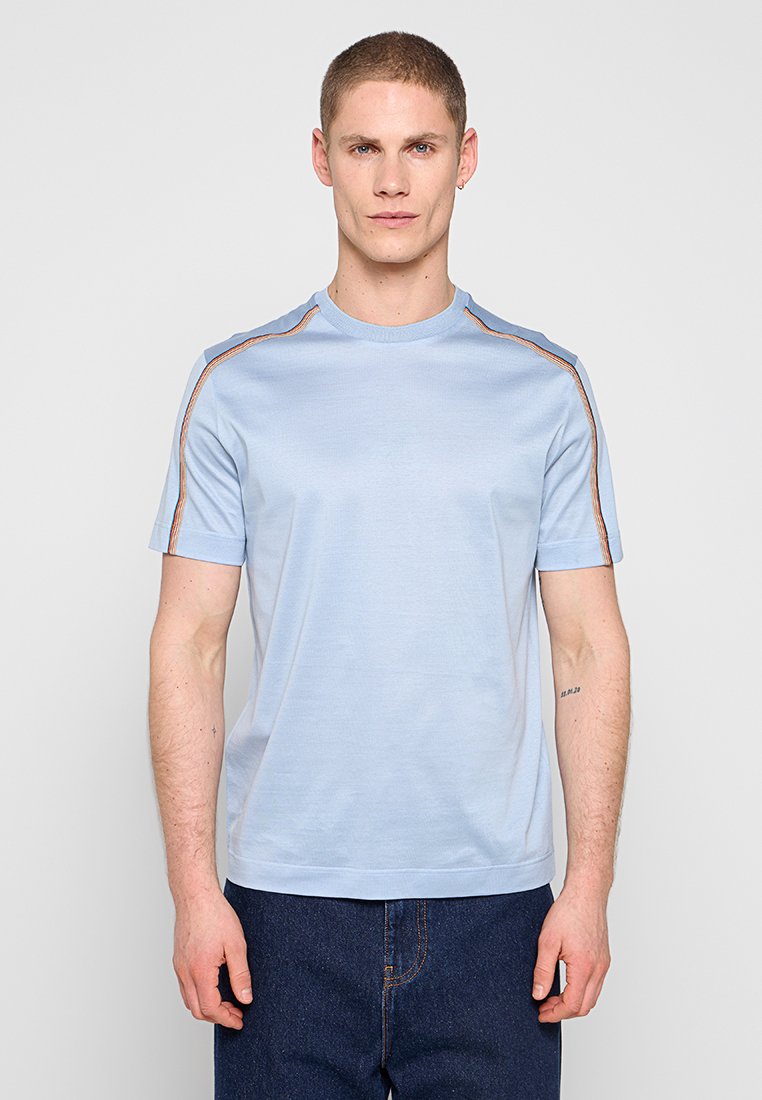 Mann mit kurzen Haaren, der ein hellblaues T-Shirt mit kurzen Ärmeln und orangefarbenen Streifen auf den Schultern sowie dunkelblaue Jeans trägt und vor grauem Hintergrund steht.