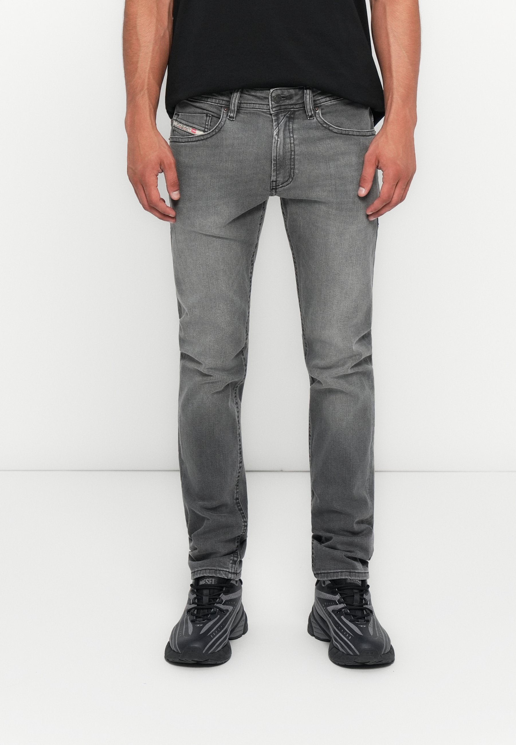 Mann in schmal geschnittenen grauen Jeans und schwarzen Sneakers vor einem weißen Hintergrund, stehend mit entspannten Händen an den Seiten.