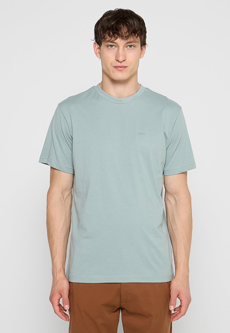 Junger Mann mit kurzen braunen Haaren, der ein hellblaues T-Shirt mit Rundhalsausschnitt und braune Hose trägt, steht vor einem einfachen hellgrauen Hintergrund.
