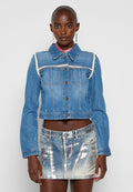 Frau trägt eine blaue, kurz geschnittene Jeansjacke mit weißem Besatz und einen metallisch silbernen sowie blauen, ausgewaschenen Jeansminirock.