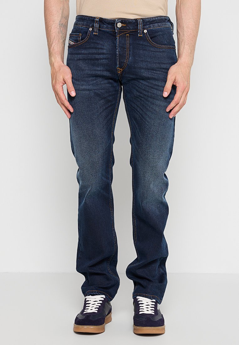 Mann trägt dunkelblaue Jeans mit geradem Bein und marineblaue Sneakers mit weißen Schnürsenkeln, steht vor einem schlichten hellen Hintergrund.
