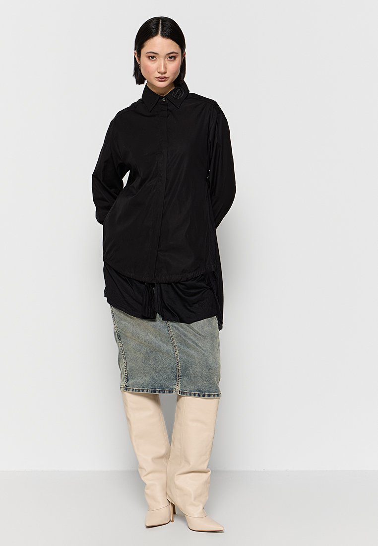 Schwarzes oversized Hemd mit Knopfverschluss vorne und abgerundetem Saum, kombiniert mit einem Jeans-Bleistiftrock und knielangen beigen Stiefeln.