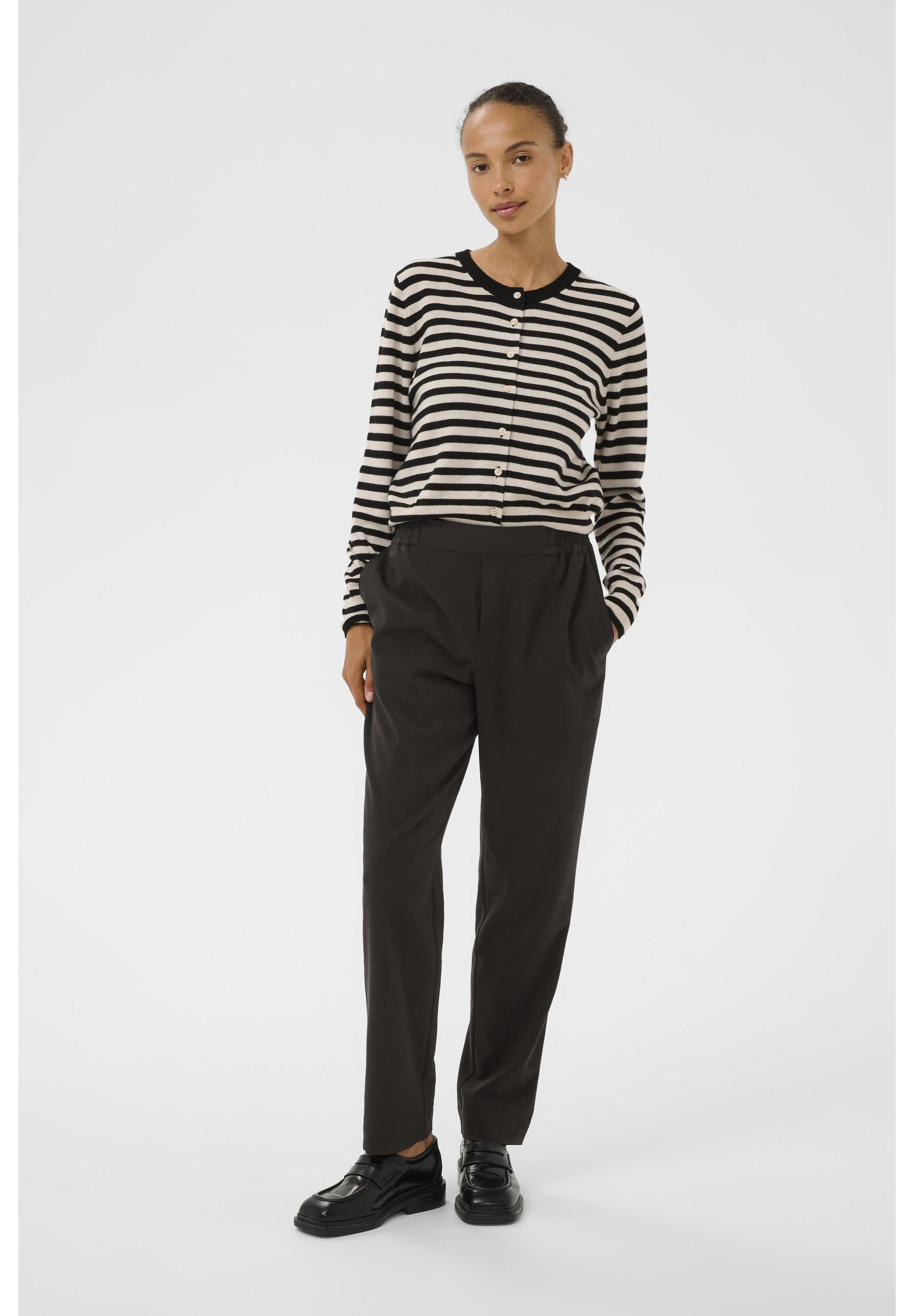Schwarz-weißer gestreifter Cardigan mit Knöpfen, kombiniert mit hoch taillierten schwarzen Hosen. Das Model trägt schwarze Schuhe und steht vor einem schlichten Hintergrund.
