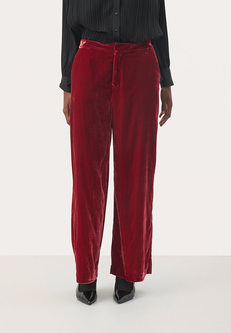 Rote Samt-Hosen mit weit geschnittenem Bein und glatter Textur, ausgestattet mit einem Kordelzugbund und einem entspannten Schnitt, kombiniert mit einer schwarzen gefältelten Bluse und schwarzen spitzen Schuhen.