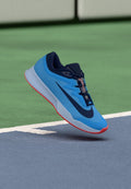 Blauer Sportschuh mit strukturiertem Design, versehen mit einem dunkelblauen Akzent und einer kontrastierenden roten Sohle, schwebend über einer Spielfläche.