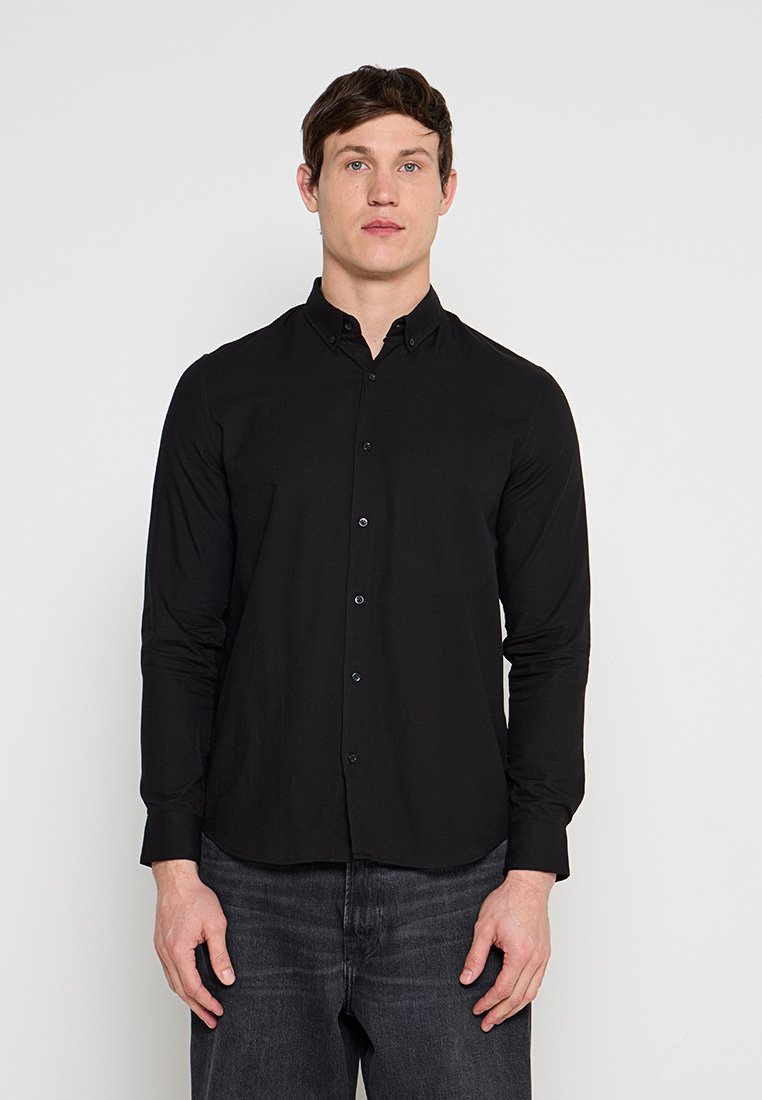 Mann trägt ein eng anliegendes langärmliges schwarzes Hemd mit Knöpfen und dunkle Jeans, steht vor einem schlichten weißen Hintergrund.