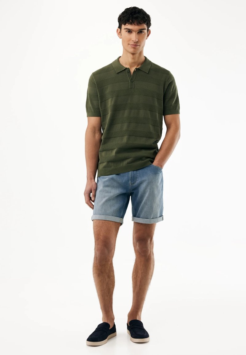 Junger Mann, der ein grünes Polohemd mit kurzen Ärmeln, hellblaue hochgekrempelte Jeansshorts und schwarze Slipper trägt.