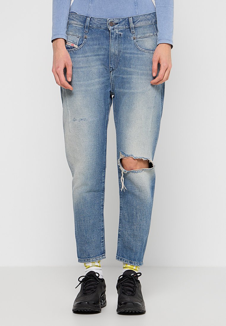 Helle blaue Denim-Jeans mit einem vernieteten Finish, featuring gerissene Knie, gerader Schnitt und klassischem Fünf-Taschen-Design. Kombiniert mit schwarzen Sneakers.