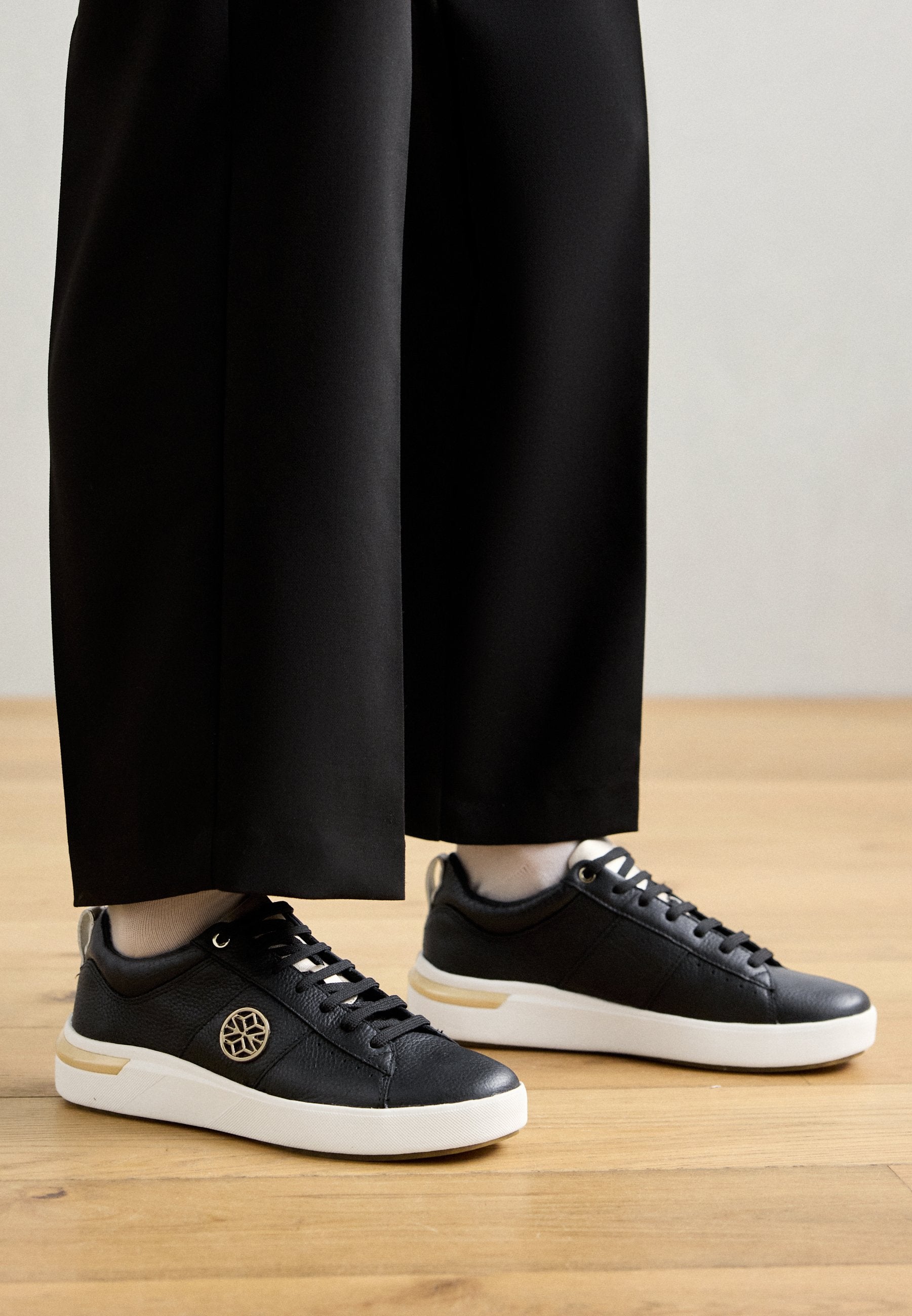 Schwarze Leder-Sneaker mit strukturiertem Finish, weißer Gummisohle und goldenen Akzenten. Die Schnürsenkel sind schwarz, mit einem runden Logo an der Seite.