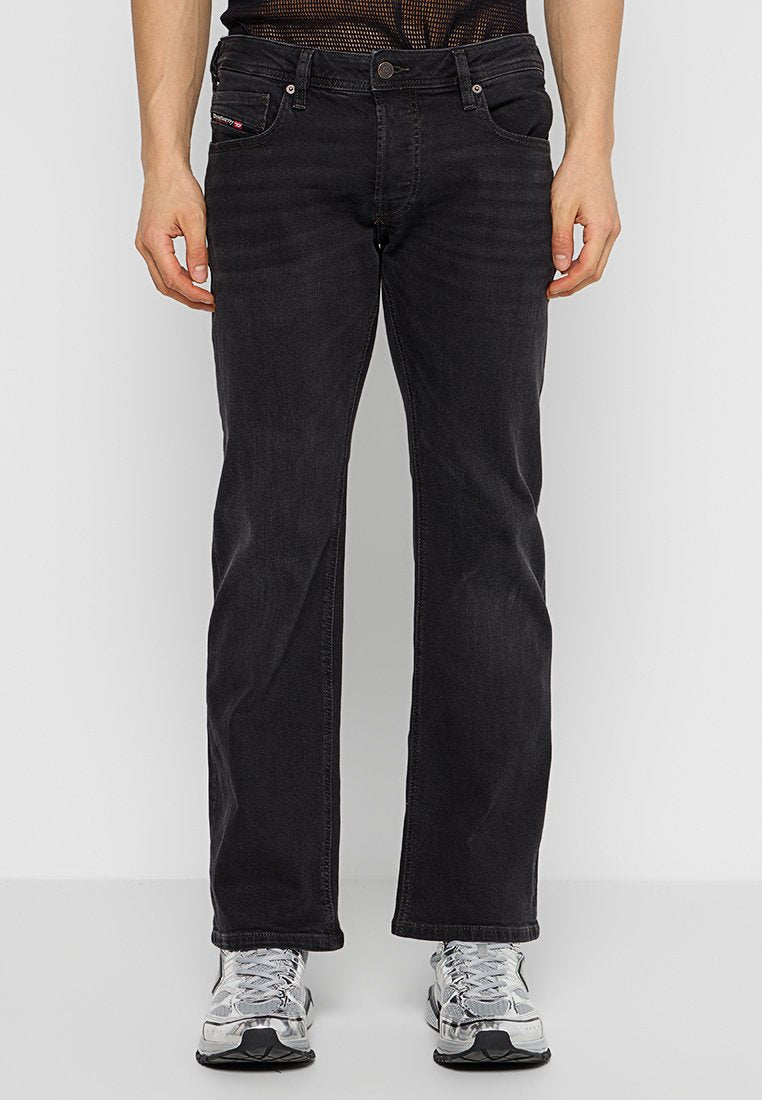 Schwarze Jeans aus Denim mit geradem Schnitt, fünf Taschen, Gürtelschlaufen und einem dezenten Logo-Tag. Getragen mit silbernen Turnschuhen und einem Netzoberteil.