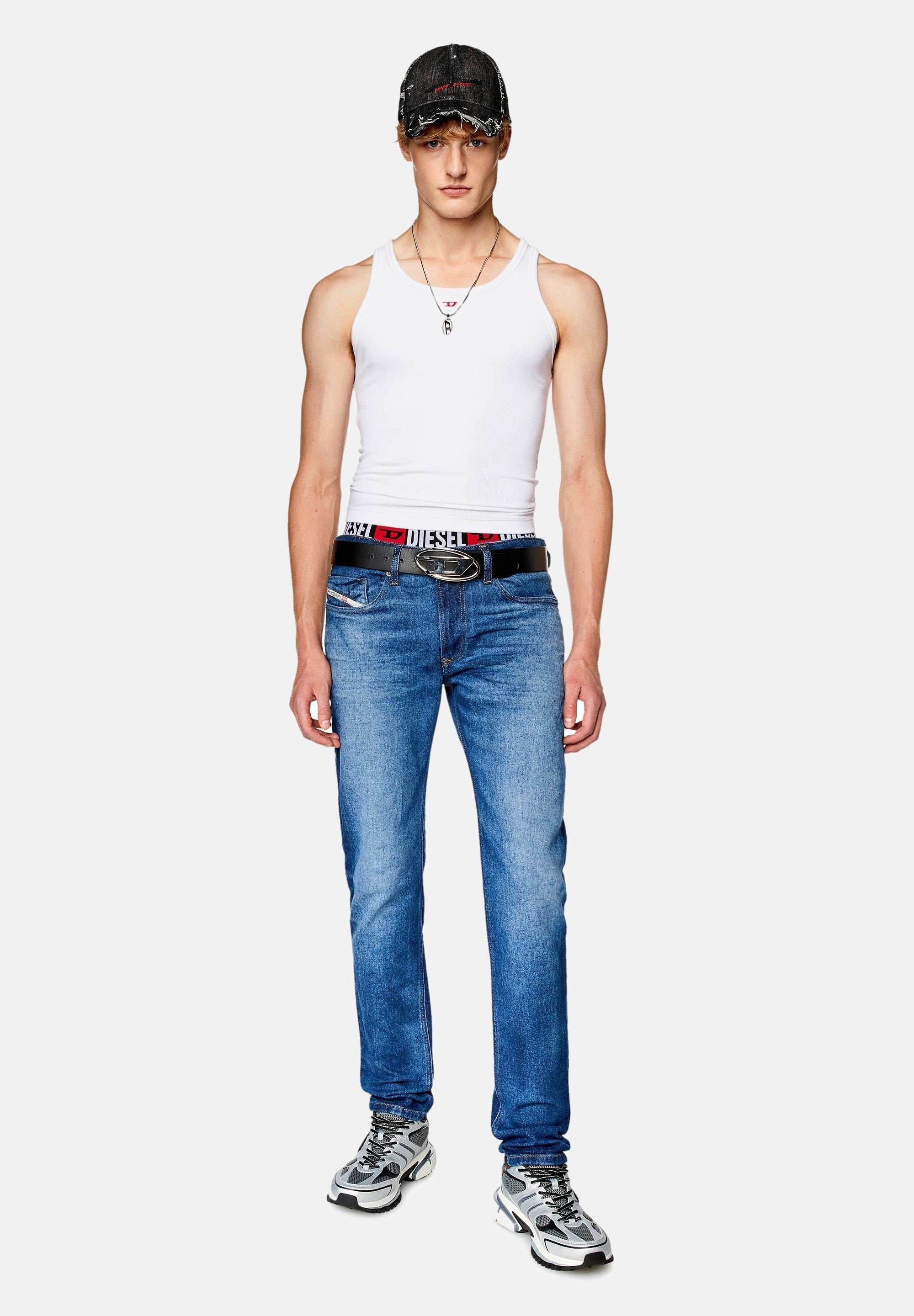 Weißes Tanktop, blaue Jeans, schwarzer Gürtel mit Logo, grau-weiße Sneakers, schwarze Mütze mit rotem Akzent. Lässiges Outfit mit modernem Design.