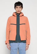 Junger Mann in einer zweifarbigen Jacke mit einem lachsfarbenen Außenstoff und einem dunkelgrünen Innenstoff, kombiniert mit hellblauen Jeans, steht vor einem weißen Hintergrund.