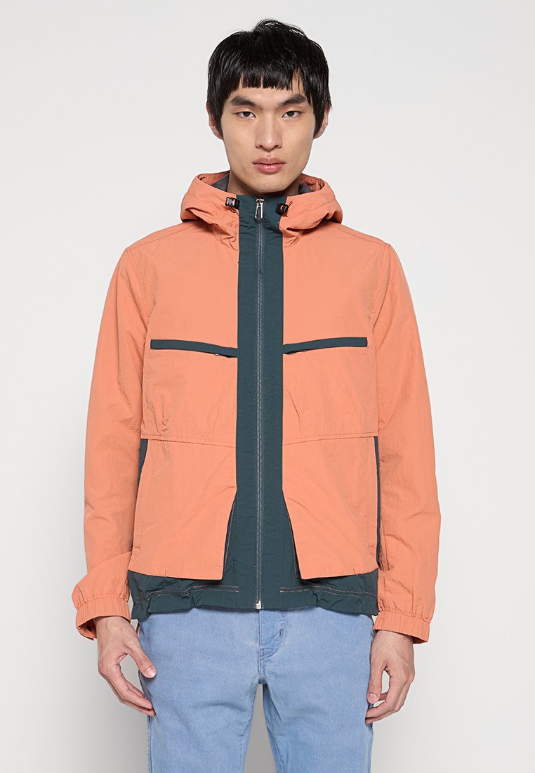 Junger Mann in einer zweifarbigen Jacke mit einem lachsfarbenen Außenstoff und einem dunkelgrünen Innenstoff, kombiniert mit hellblauen Jeans, steht vor einem weißen Hintergrund.