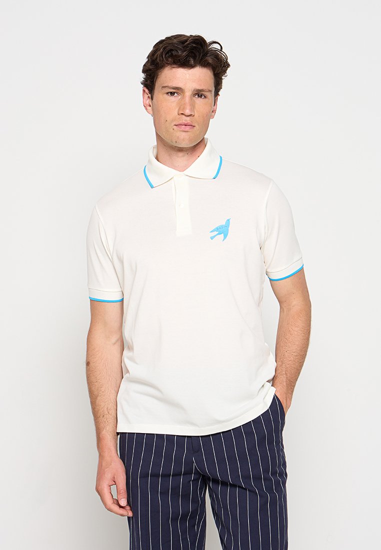 Mann trägt ein weißes Poloshirt mit blauen Akzenten und Vogel-Logo, kombiniert mit marineblauen Nadelstreifenhosen, steht vor einem schlichten Hintergrund.