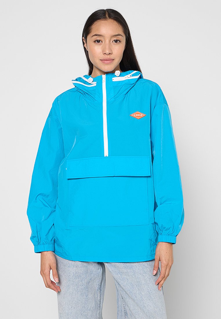 Helle blaue wasserdichte Jacke mit einer Fronttasche, verstellbarem Kapuze und einem weißen Reißverschluss; mit orangefarbenem Logodetail auf der Brust.