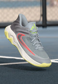 Graue und gelbe Sporttrainer mit rotem Swoosh-Detail, schwebend über einer blauen Außen-Basketballplatzoberfläche in der Nähe eines Maschendrahtzauns.