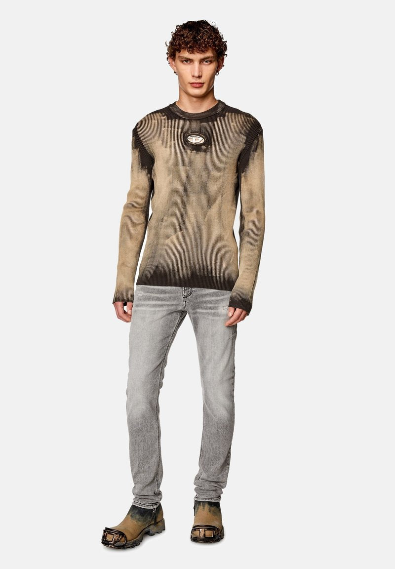 Strickpullover in Brauntönen und Schwarz mit gerippter Textur, versehen mit einem runden Logodetail auf der Brust, kombiniert mit hellgrauen Jeans und schwarzen Stiefeln.
