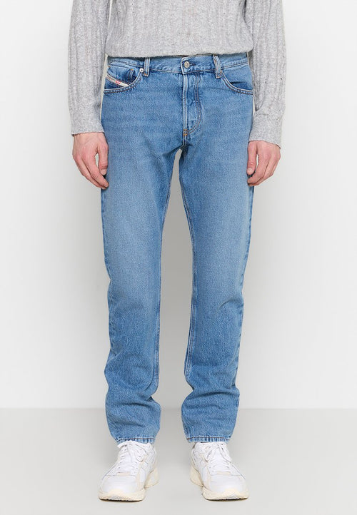 Person in hellblauen Jeans, weißen Sneakern und einem grauen Strickpullover, der vor einem einfarbigen hellen Hintergrund steht.