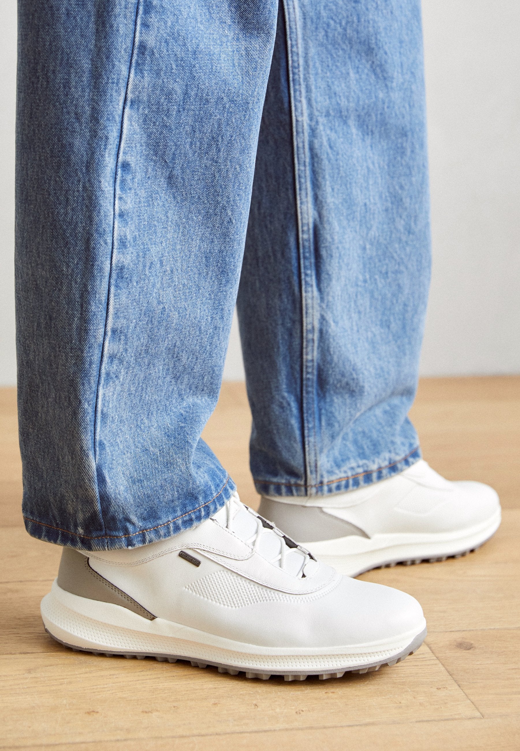 Weiße Sportschuhe mit strukturiertem Material und grauen Akzenten, kombiniert mit locker sitzenden blauen Jeans. Sichtbare Gummisohle und Schnürdetails.