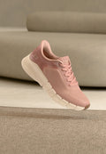 Hellrosa sportlicher Sneaker mit weißer, segmentierter Sohle, Mesh-Obermaterial und Schnürsenkeln, auf einem beigen Hintergrund mit Teppich und Sofa.