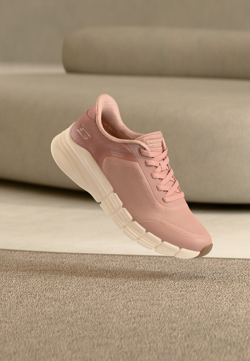 Hellrosa sportlicher Sneaker mit weißer, segmentierter Sohle, Mesh-Obermaterial und Schnürsenkeln, auf einem beigen Hintergrund mit Teppich und Sofa.