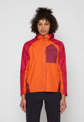 Frau mit lockigem Haar, die eine rot-orange Outdoor-Jacke mit Reißverschluss und schwarze Hose trägt, steht vor einem einfarbigen hellgrauen Hintergrund.