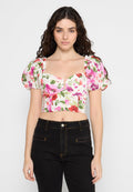 Blumiges Crop-Top mit Puffärmeln, bestehend aus einem weißen Grund mit rosa, roten und grünen Mustern sowie einem zentralen Knopfdetail. Kombiniert mit schwarzen Jeans.