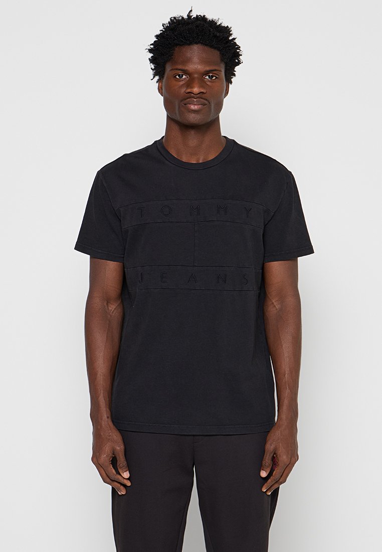 Mann mit dunkler Haut und kurzen lockigen Haaren, der ein schwarzes Tommy Jeans T-Shirt und schwarze Hosen trägt und vor einem schlichten hellen Hintergrund steht.