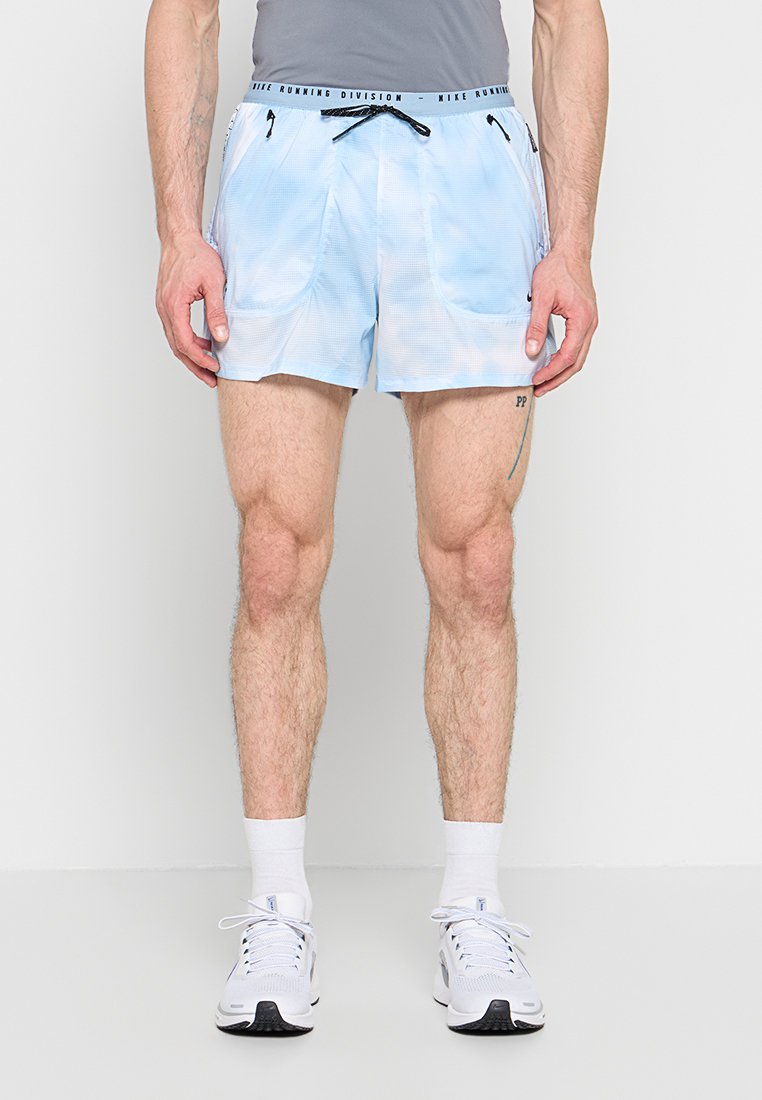 Mann trägt hellblaue Nike-Laufshorts mit Reißverschlusstaschen, weiße Socken und weiße Sportschuhe, steht vor einem schlichten Hintergrund.