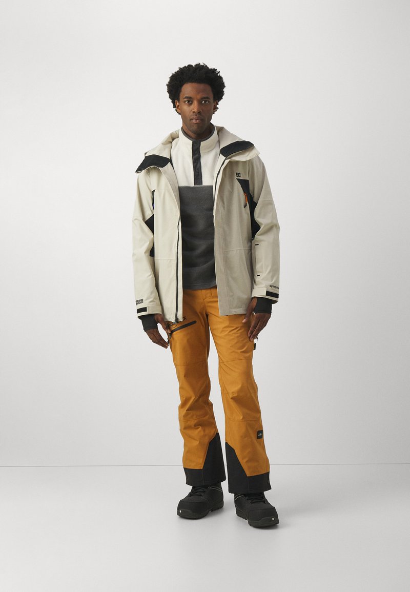 Wasserdichte, hellgraue Jacke mit schwarzen Akzenten, grauem Midlayer und orangefarbenen Skihosen. Verfügt über Reißverschlüsse und eine Kapuze. Schwarze Schuhe.