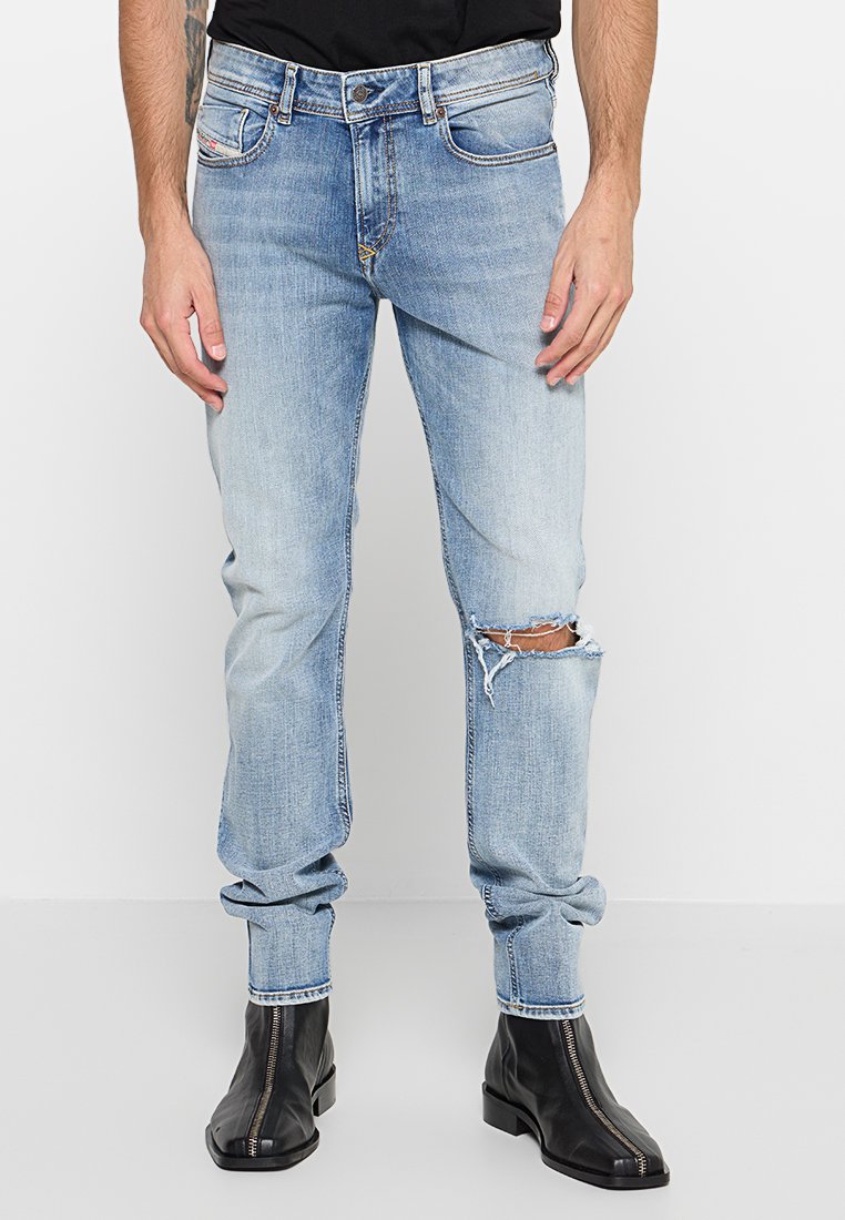 Hellblaue Jeans im Slim-Fit-Schnitt, mit distressed Knie-Öffnungen und einer leicht verwaschenen Textur. Kombiniert mit schwarzen Stiefeletten.