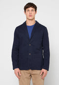 Marineblauer Baumwollblazer mit zwei vorderen Taschen, aufgeschlagenen Revers und zwei braunen Knöpfen. Getragen über einem blauen Rundhalsausschnitt-Shirt.