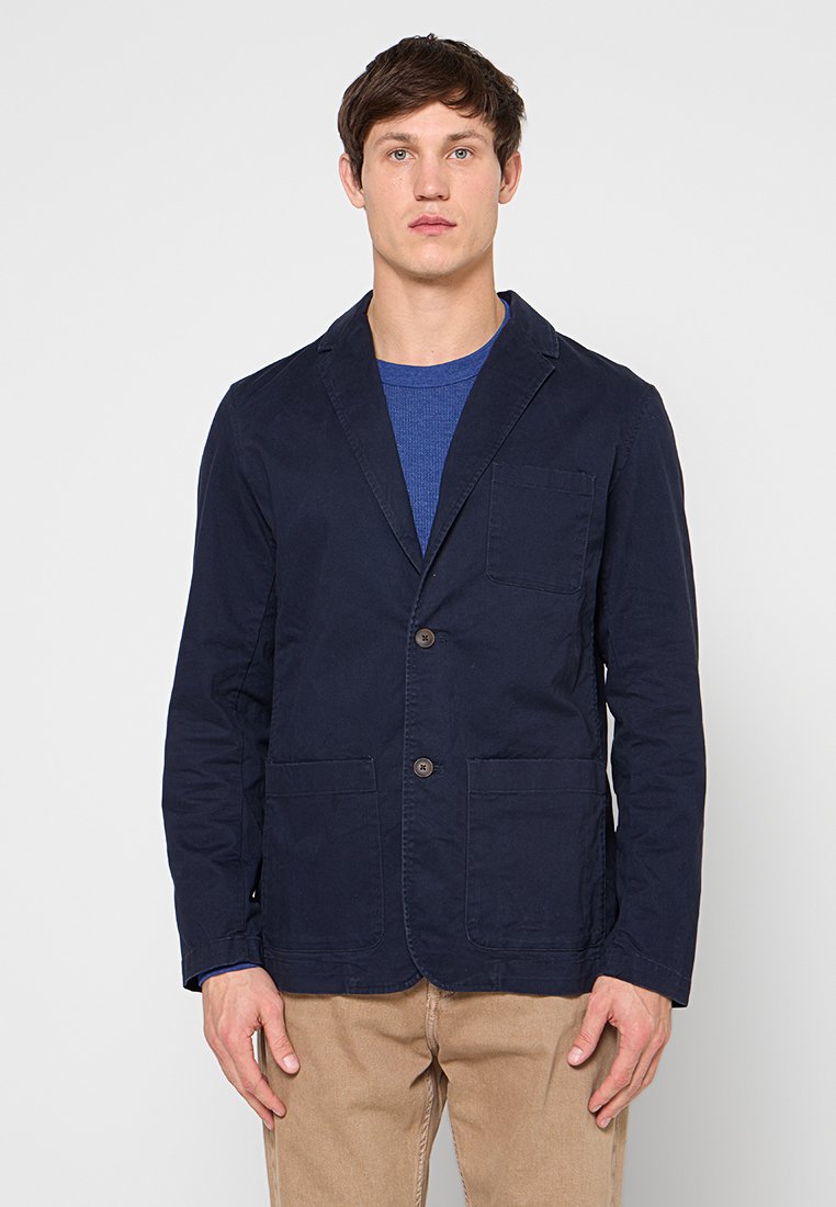 Marineblauer Baumwollblazer mit zwei vorderen Taschen, aufgeschlagenen Revers und zwei braunen Knöpfen. Getragen über einem blauen Rundhalsausschnitt-Shirt.