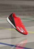Roter und schwarzer Hallenschuh mit orangen Schnürsenkeln, weißer Sohle und großem weißem Logo, das über dem Hallenboden mit bunten Spielfeldlinien schwebt.