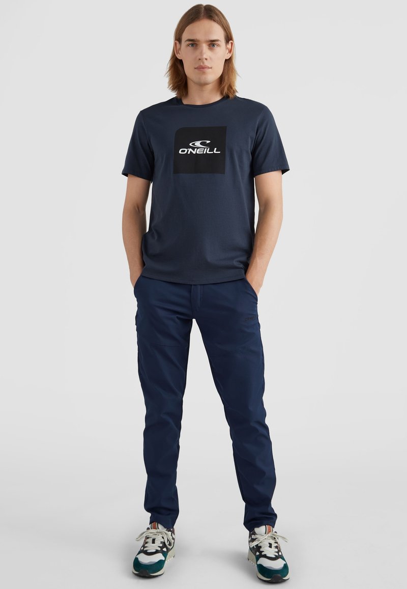 Dunkelblaues T-Shirt mit großem weißem Logo auf der Vorderseite, kombiniert mit dunkelblauen Hosen und mehrfarbigen Turnschuhen. Glatte Stofftextur.