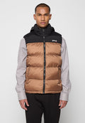 Weste aus Steppmaterial mit der oberen Hälfte aus schwarzem Nylon und der unteren Hälfte in braunem Steppmuster. Mit einem Reißverschluss und einem Logo auf der Brust. Getragen über einem gestreiften Hemd.