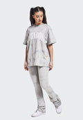 Junge Frau mit langen, dunklen Haaren in Zöpfen, die ein graues Batik-Oversize-T-Shirt und passende Hosen trägt, dazu weiße Sneakers und nach unten schaut.