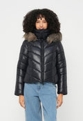 Frau mit lockigem Haar, die eine schwarze gesteppten Jacke mit Kapuze aus Fell und hellblauen Jeans trägt, steht vor einem schlichten Hintergrund.