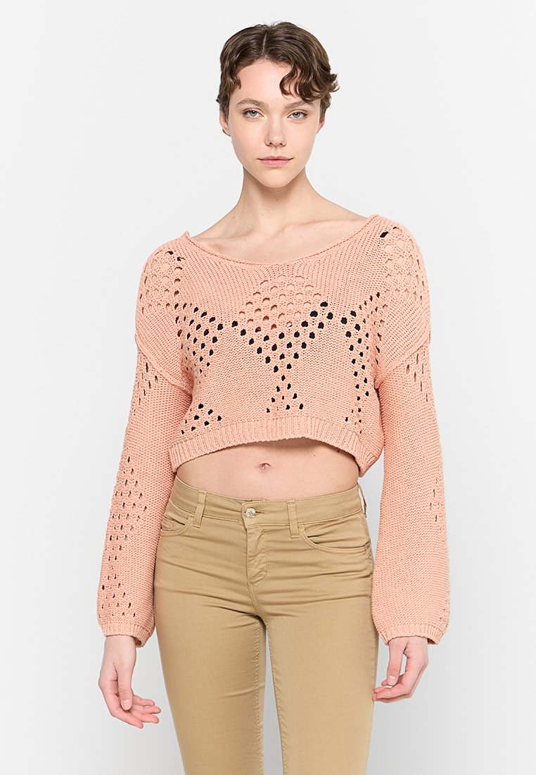 Rosa, gestrickter, cropped Pullover mit offenen Netzmustern und weiten Ärmeln. Kombiniert mit hellbeigen, figurbetonten Hosen.