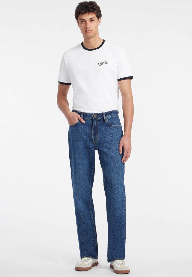 Weißes Baumwoll-T-Shirt mit schwarzem Rand und "Guess"-Logo, kombiniert mit blauen Jeans, die eine lässige Passform und klassisches Design zeigen.
