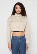 Frau mit lockigem Haar, die eine beige, kurz geschnittene Jacke mit Knöpfen und hoch taillierte blaue Jeans trägt, steht vor einem schlichten Hintergrund.